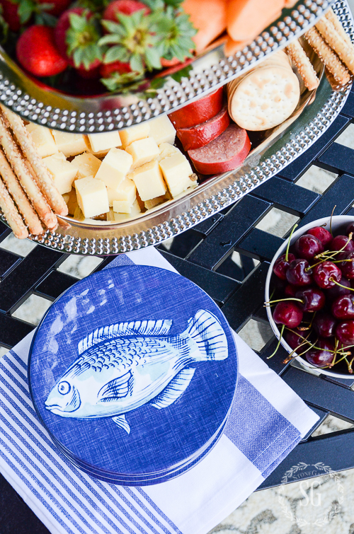 Blue fish melamine plates with striped napkin, silver appetizer tray, and bowl of cherries on a black iron patio table for summer al fresco dining