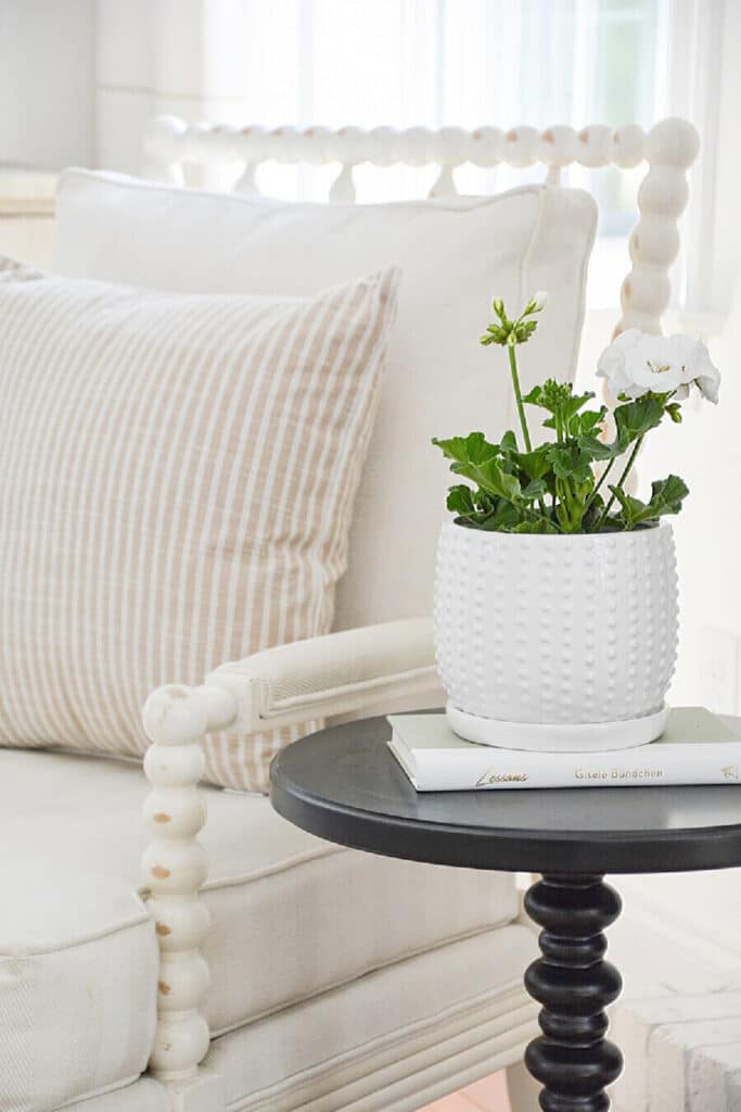 a white pot with a white geranium in it sitting on a drink table beside a white chair.