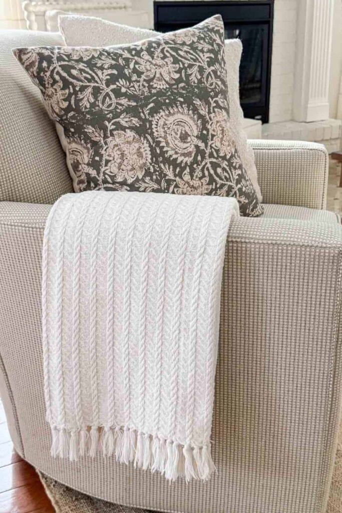 White cable knit throw with fringe draped over a neutral upholstered chair with a dark floral pillow