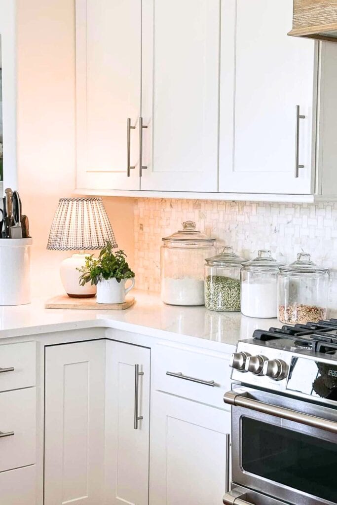 organized kitchen counter with glass pantry canisters, small lamp, and white cabinets