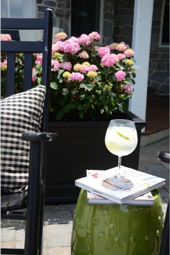 pink hydrangeas overflowing a black rectangular planter on a stone patio with a black rocking chair, gingham pillow, and a cool drink on a green garden stool