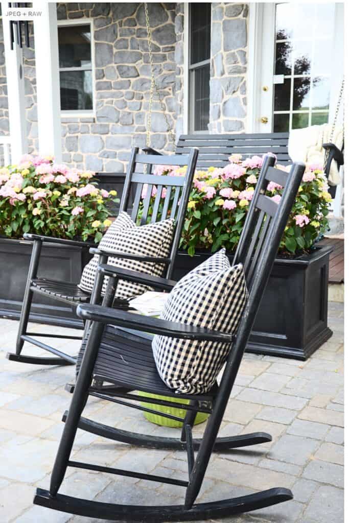 black rocking chairs with black and white gingham pillows on a stone patio with large planters of pink hydrangeas against a stone house