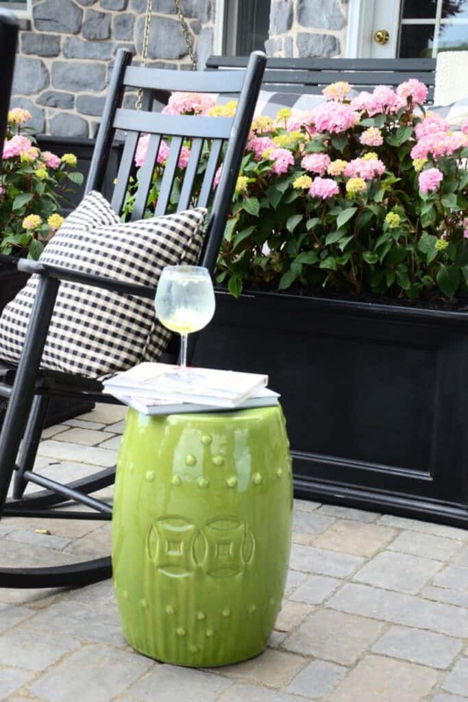 black rocking chair with black and white gingham pillow next to a lime green garden stool with a wine glass and magazines against a backdrop of pink hydrangeas on a summer patio