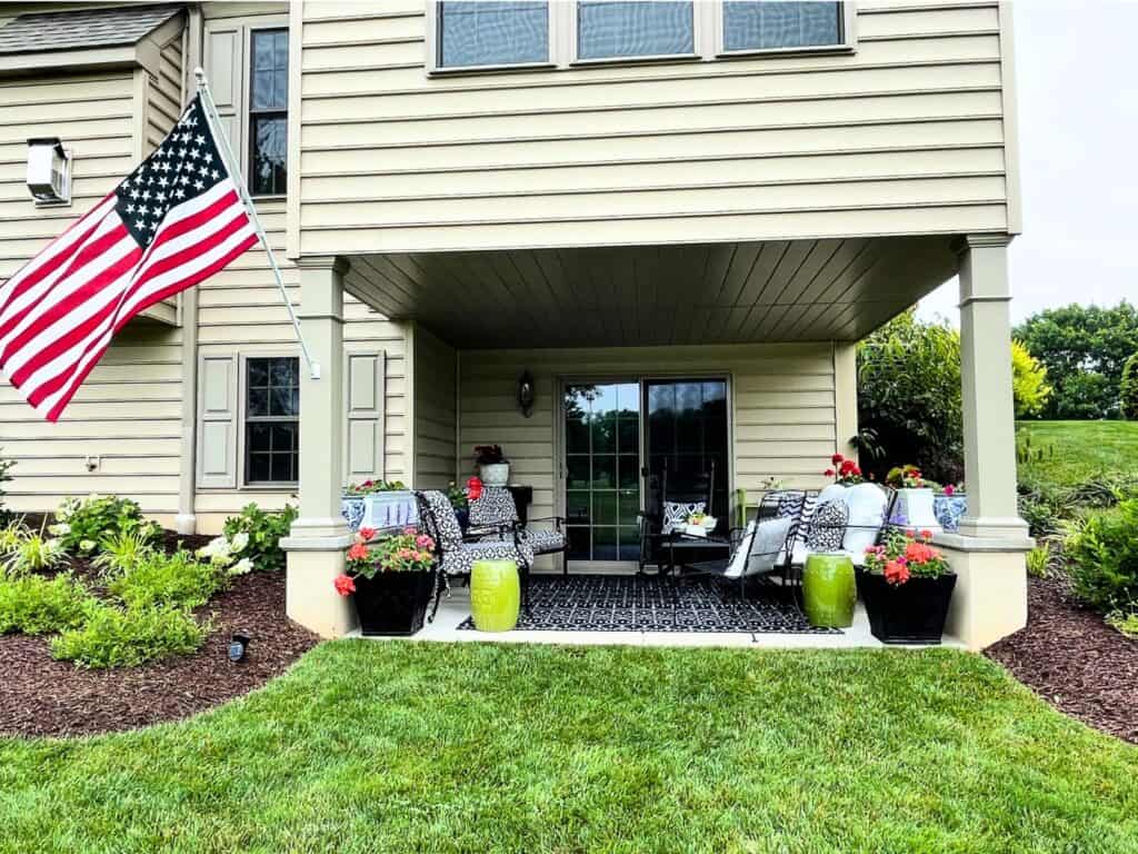 covered patio with black and white patterned outdoor furniture, lime green garden stools, black planters with red geraniums, and an American flag on a summer day
