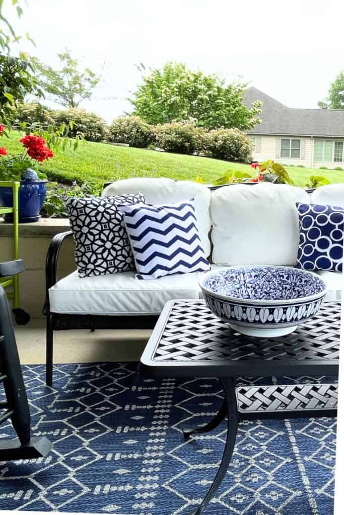 black metal outdoor sofa with white cushions and navy blue and white patterned throw pillows on a summer patio with a blue and white chinoiserie bowl on the coffee table