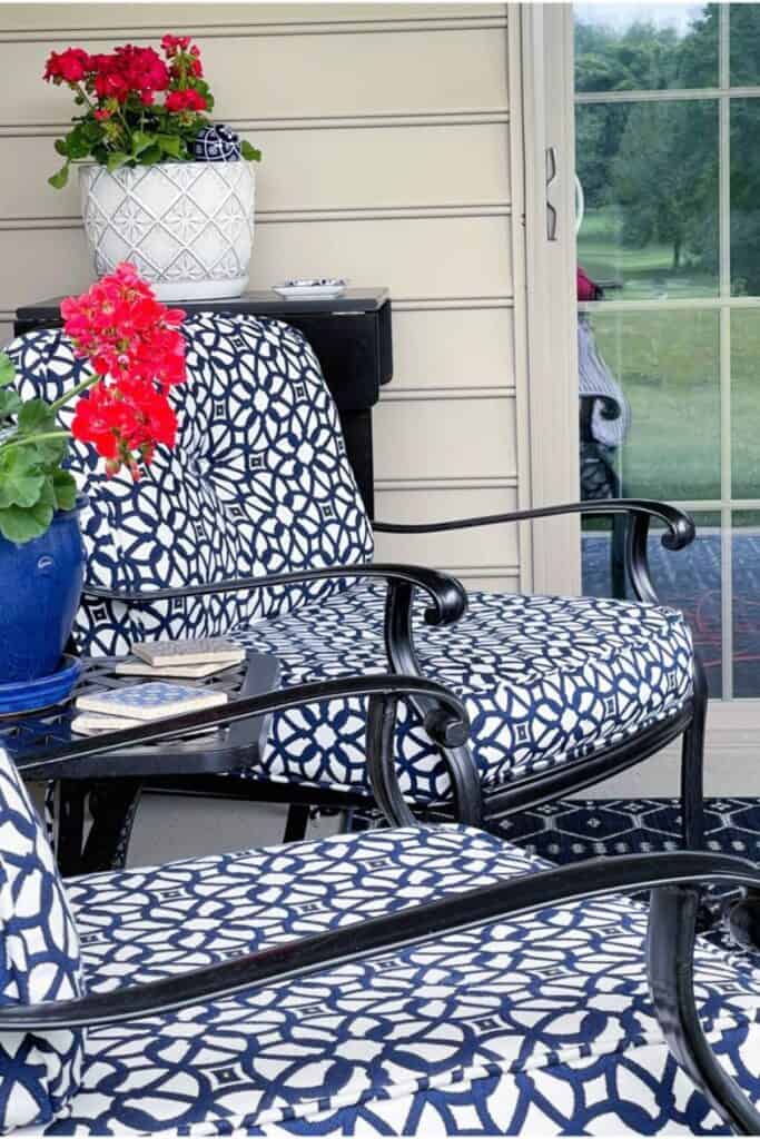 black cast iron patio chairs with navy blue and white patterned cushions, red geraniums in a blue pot, and a white textured planter on a covered summer patio