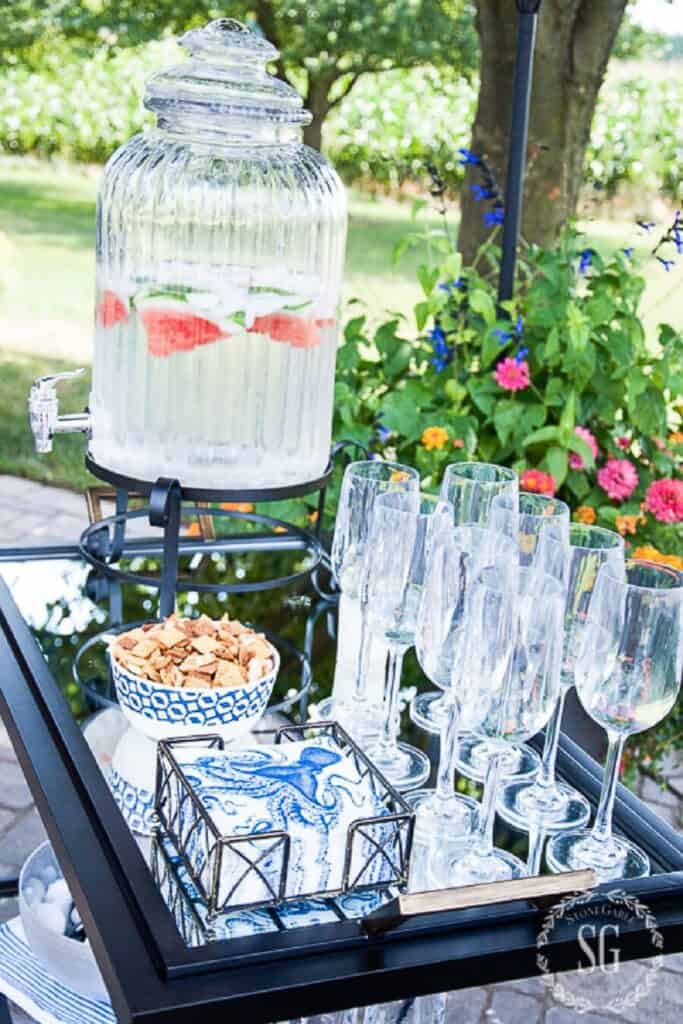 outdoor drink station with glass beverage dispenser filled with watermelon water, champagne flutes, and blue and white accessories on a summer patio