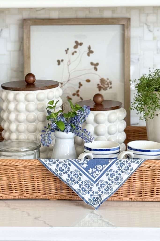 simple kitchen ideas using a wicker tray with blue and white decor, canisters, mugs, and flowers on a kitchen countertop