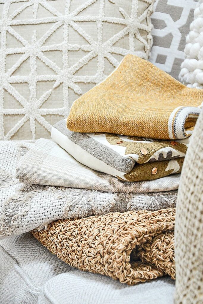 Stack of textured throw pillows and folded throws in neutral and mustard tones for late summer decorating