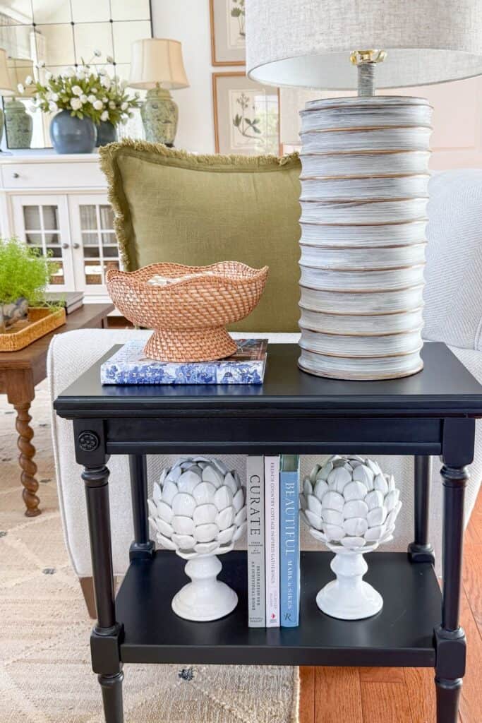 Black side table styled with a ribbed lamp, woven basket, olive pillow, and white ceramic artichoke bookends
