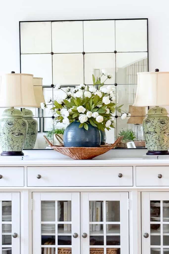 Blue ceramic vase with white florals in a woven basket on a white buffet flanked by green chinoiserie lamps