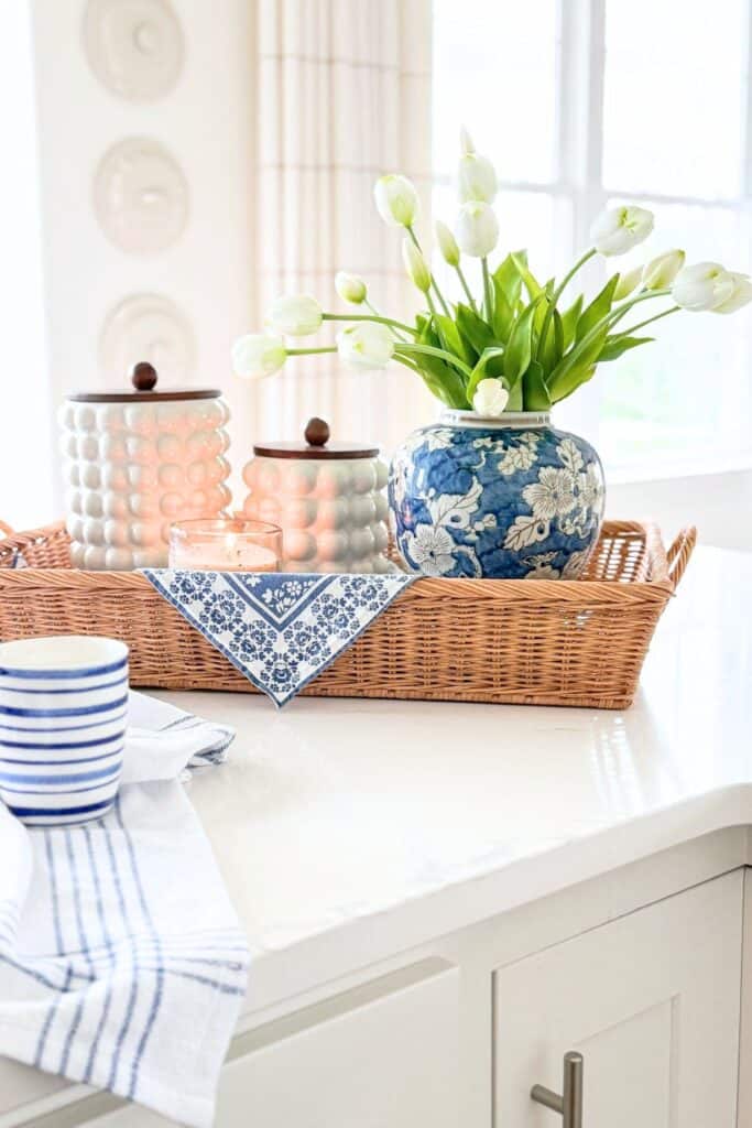 Blue and white chinoiserie vase with white tulips styled in a woven tray with hobnail canisters on a kitchen island