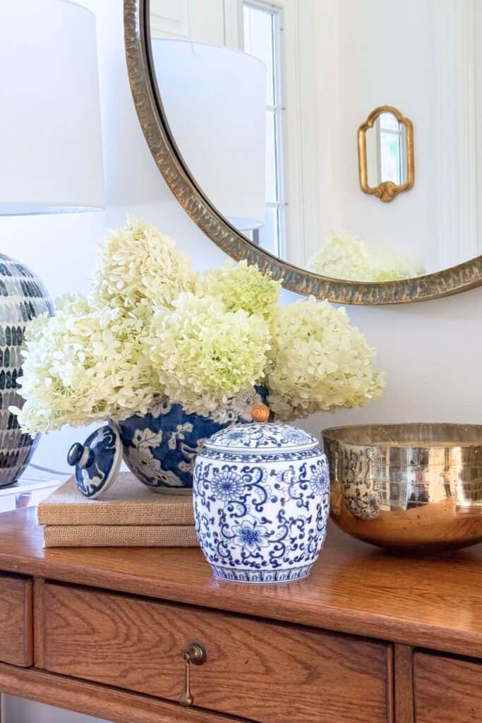 Blue and white ginger jars with hydrangeas styled beside a brass bowl under a round mirror on a wood console
