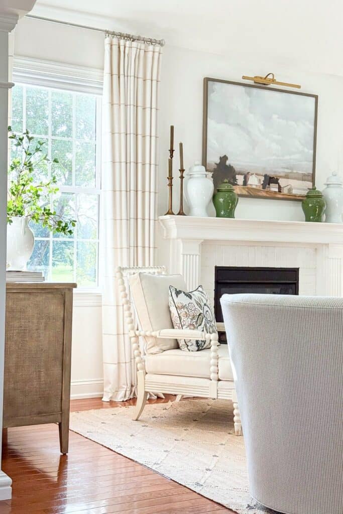 Living room corner with a white mantel styled with green ginger jars and brass candlesticks beside a cream spool chair