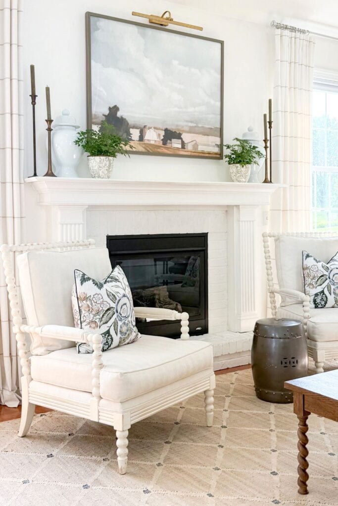 White fireplace mantel styled with brass candlesticks and landscape art flanked by cream spool chairs with floral pillows