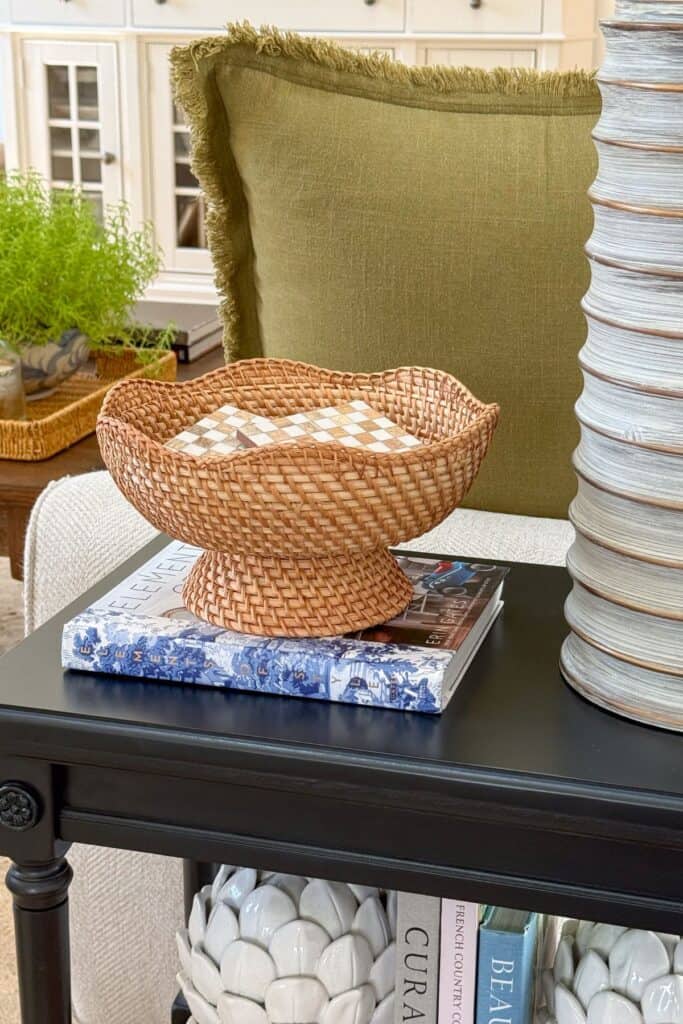 Woven pedestal basket with a checkerboard box styled on a black table beside an olive linen pillow and ribbed lamp base