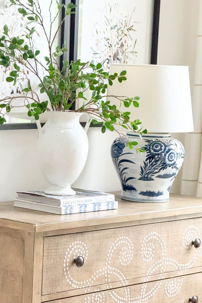 A dresser top styled with stacked books used as risers under a white vase filled with fresh green branches, alongside a blue and white ginger jar lamp, showing how to decorate with what you already have.