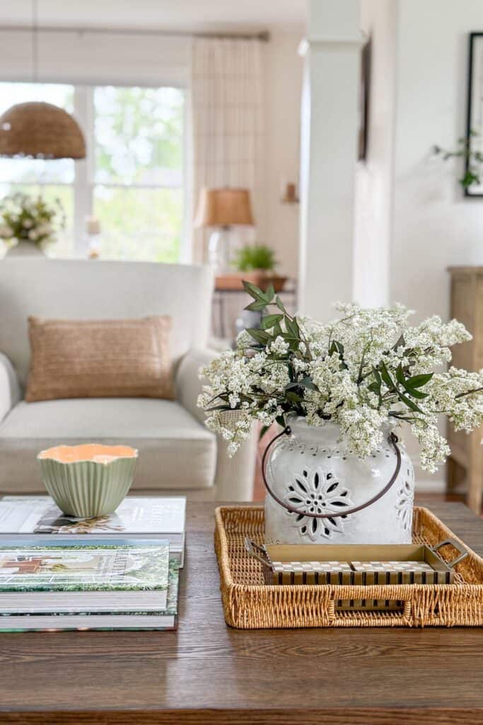 A wood coffee table styled with a wicker tray holding a white ceramic vase filled with fresh white clippings and greenery, stacked books, and a small green bowl at Tanglewood House showing how to decorate with what you already have.