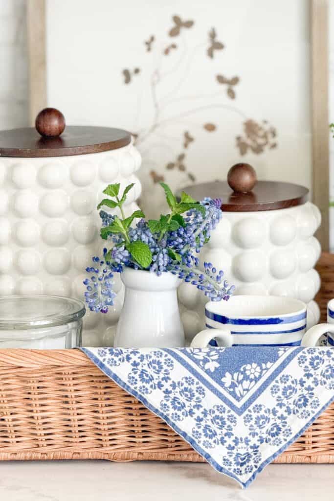 A kitchen counter vignette at Tanglewood House styled with white hobnail canisters, blue and white mugs, a small white pitcher with fresh purple clippings and mint, a blue and white floral napkin, and a wicker tray showing how to use kitchen items as decor.