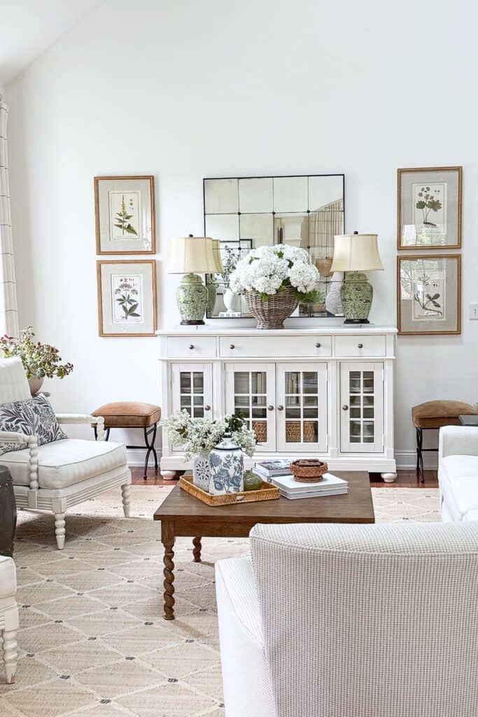 Neutral living room at Tanglewood House with a white sideboard styled with green ginger jar lamps, a wicker basket of white hydrangeas, botanical prints, and a coffee table tray with books and a blue and white ginger jar.