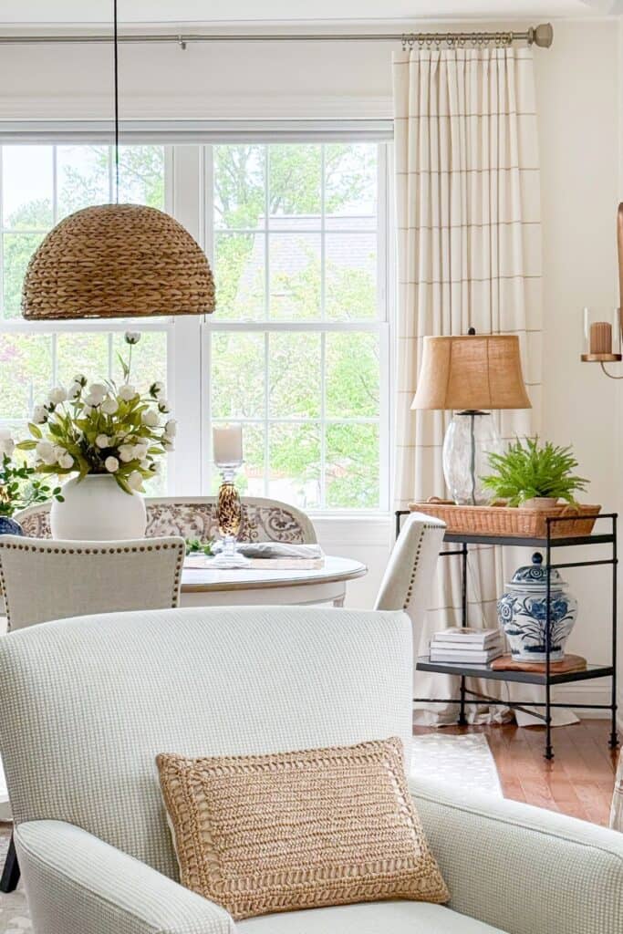 An open concept living and dining room at Tanglewood House showing a styled bar cart with a wicker tray, blue and white ginger jar, and stacked books alongside a dining table centerpiece of white blooms in a round white vase with candles.