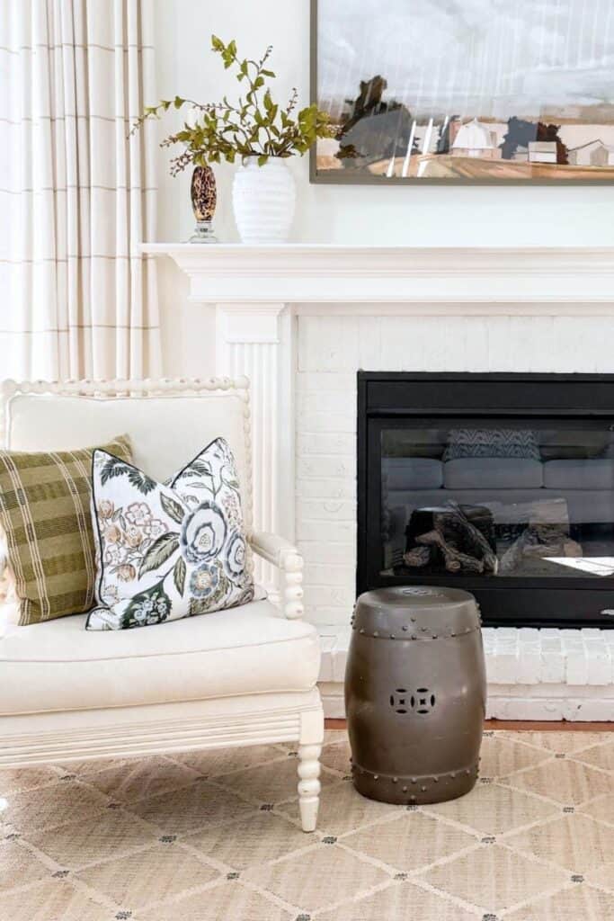 Repainted dark garden stool beside a white spindle chair with mismatched floral and plaid pillows next to a fireplace, showing how to give what you already own a fresh new look.