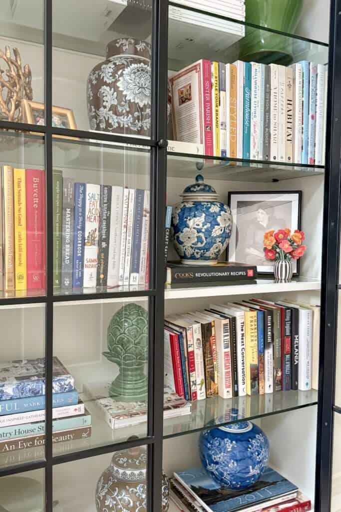 A glass door bookcase styled with a mix of vertical and horizontal books, brown and blue and white ginger jars, a green artichoke finial, framed art, and small decor pieces showing how to style a bookshelf with what you already own.