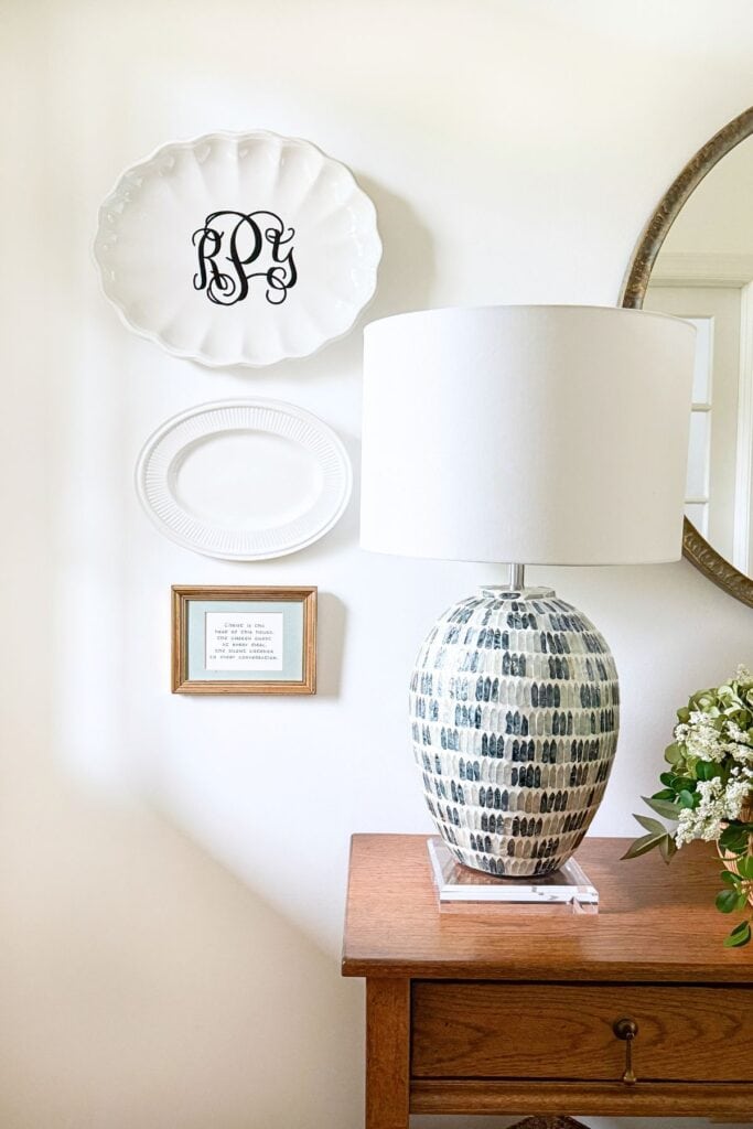 White monogrammed platter and oval dish hung on a wall above a wood dresser styled with a patterned lamp on an acrylic riser and fresh greenery, showing how to use kitchen dishes as wall decor.