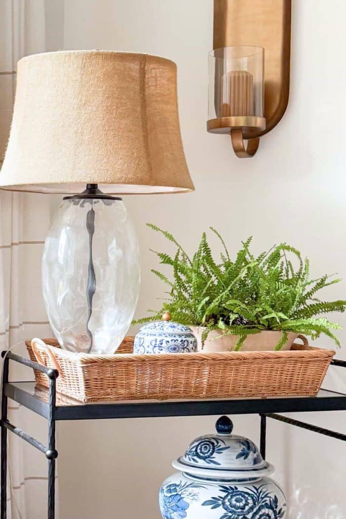A wicker basket tray styled on a black bar cart with a glass lamp, small blue and white ginger jar, and a lush fern in a terracotta pot, showing how to use trays to style and organize home decor.