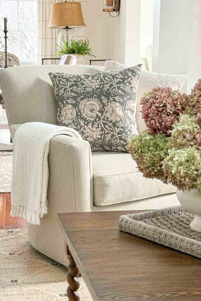 neutral slipcovered chair with a floral pillow and cream throw blanket next to a woven tray with a white bowl of pink and green hydrangeas on a wood coffee table