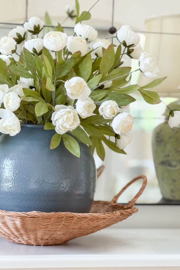 Matte blue vase with white paper florals styled in a woven basket on a mantel with a glossy green vase in the background