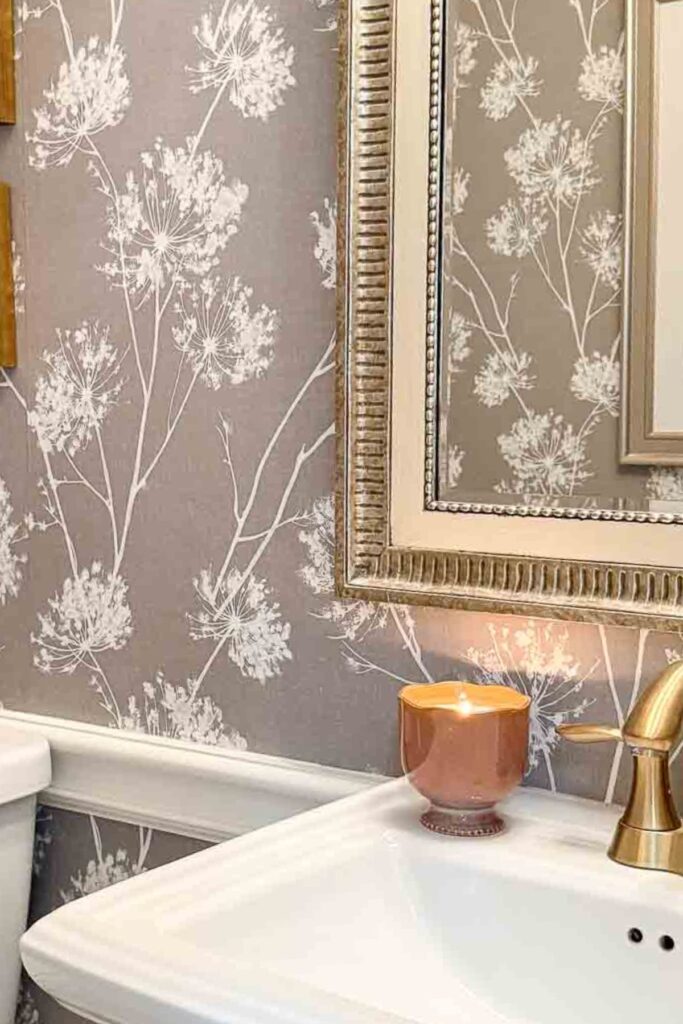 powder room with gray and white botanical wallpaper, a gold framed mirror, lit copper candle, and brass faucet on a white pedestal sink