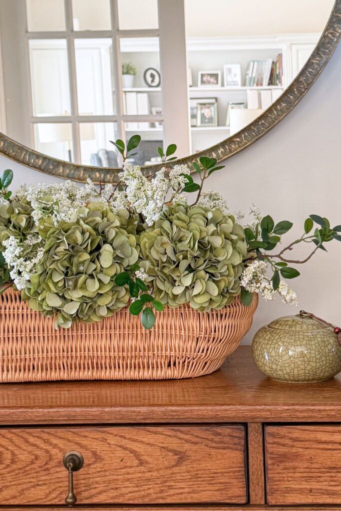 wicker basket filled with green faux hydrangeas and white florals on a wood dresser beneath a large round antiqued mirror with a small green chinoiserie ginger jar alongside