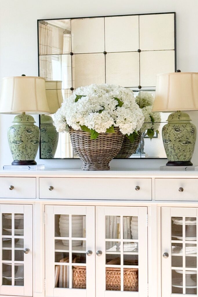neutral slipcovered chair with a floral pillow and cream throw blanket next to a woven tray with a white bowl of pink and green hydrangeas on a wood coffee table