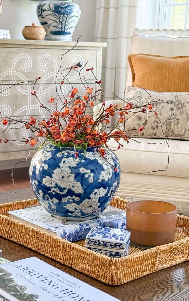 Fall coffee table decor with orange bittersweet in a blue and white jar, an amber candle, and books in a rattan tray on a dark wood coffee table