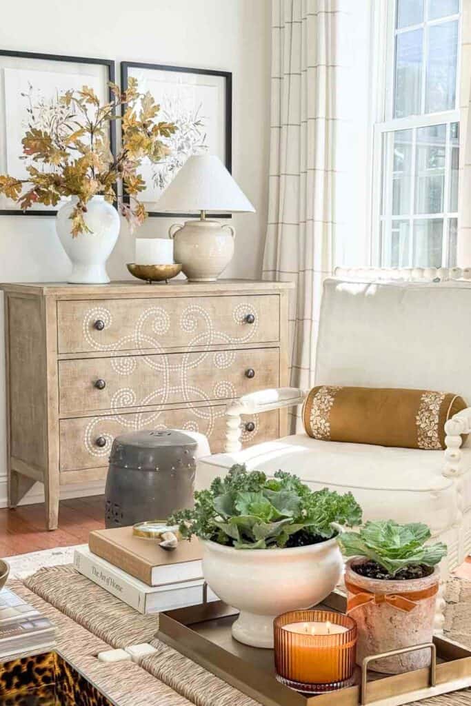 Neutral fall living room with a white armchair, natural wood chest with decorative inlay, potted ornamental cabbages, autumn branches in a vase, and a glowing candle.