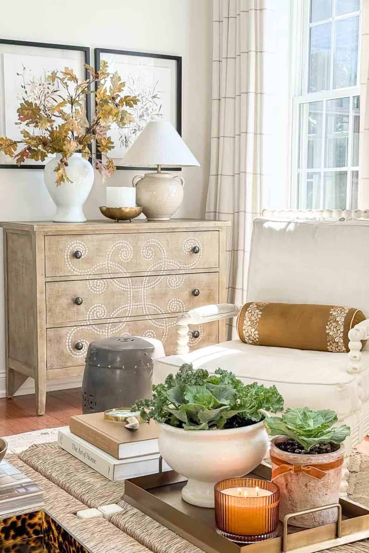 Neutral fall living room with a white armchair, natural wood chest with decorative inlay, potted ornamental cabbages, autumn branches in a vase, and a glowing candle.