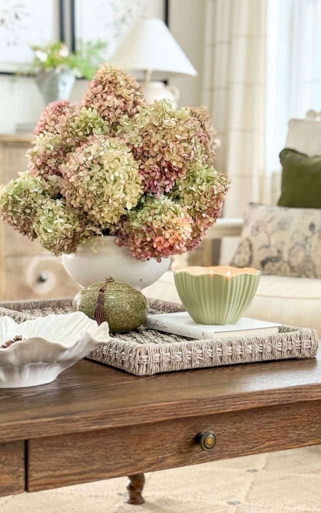 Fall vignette on a wood coffee table with green and dusty pink hydrangeas in a white bowl, a sage green candle, and a crackle glaze jar on a woven tray