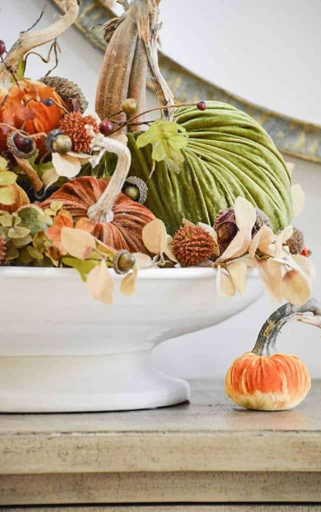 Fall vignette in earthy tones with orange, rust, and green velvet pumpkins, acorns, and berries in a white footed bowl
