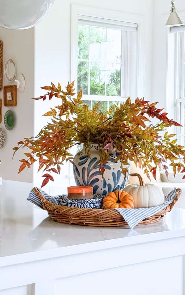 Fall vignette on a kitchen counter with autumn leaf stems in a blue and white vase, a candle on stacked bowls, and two pumpkins on a wicker tray with striped linen