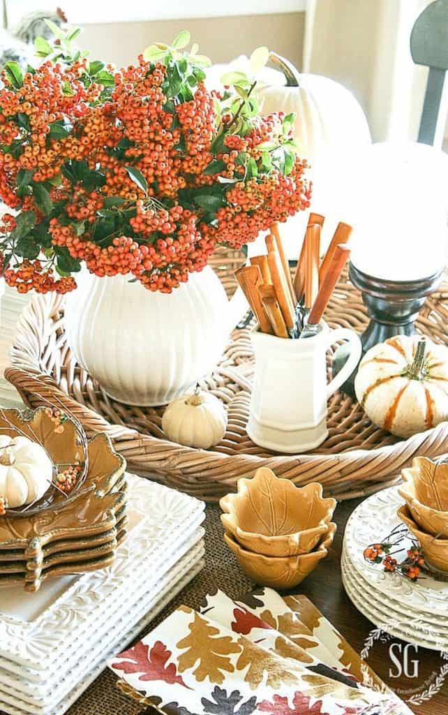 Fall vignette on a dining table with orange berry stems in a white pitcher, mini white and striped pumpkins, cinnamon sticks, and leaf shaped bowls on a woven tray