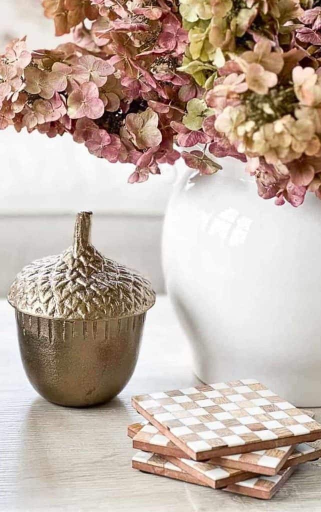 Fall vignette without a basket with dried pink hydrangeas in a white vase, a brass acorn box, and stacked checkered coasters on a table