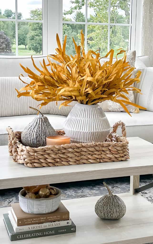 Fall vignette on a two tier coffee table with golden fall stems in a white ribbed vase, a gray gourd, a candle, and a bowl of small gourds on stacked books