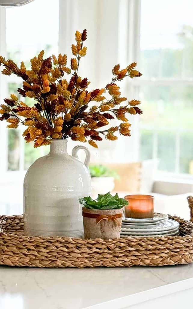 Fall vignette on a counter with golden fall stems in a white jug, a small potted plant, and a candle on stacked plates on a woven tray