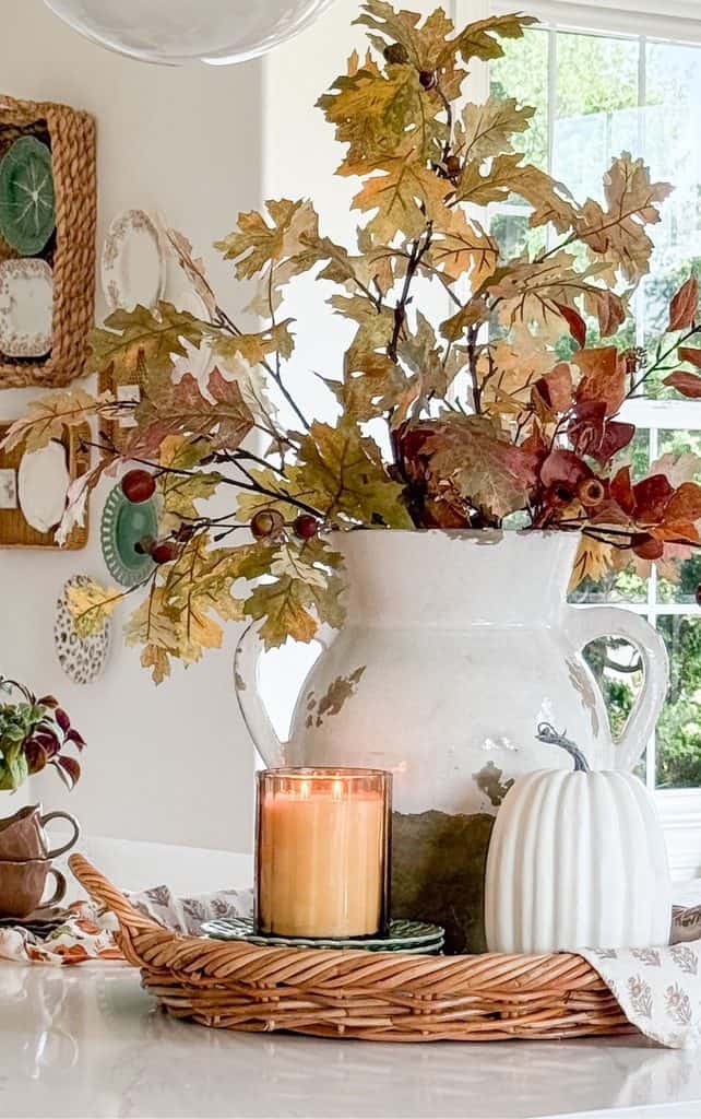 Fall vignette on a kitchen counter with oak leaf stems in a white crackled pitcher, a lit candle, and a white ribbed pumpkin on a wicker tray