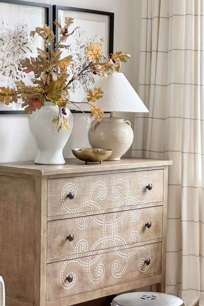 Rustic wooden dresser with decorative white dotted swirl pattern, vintage knob hardware, topped with autumnal decor and a ceramic table lamp, in a cozy living space.