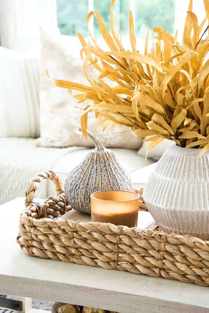 Budget friendly fall vignette in a woven basket with a candle, textured pumpkin, and golden leaves in a white vase on a coffee table.