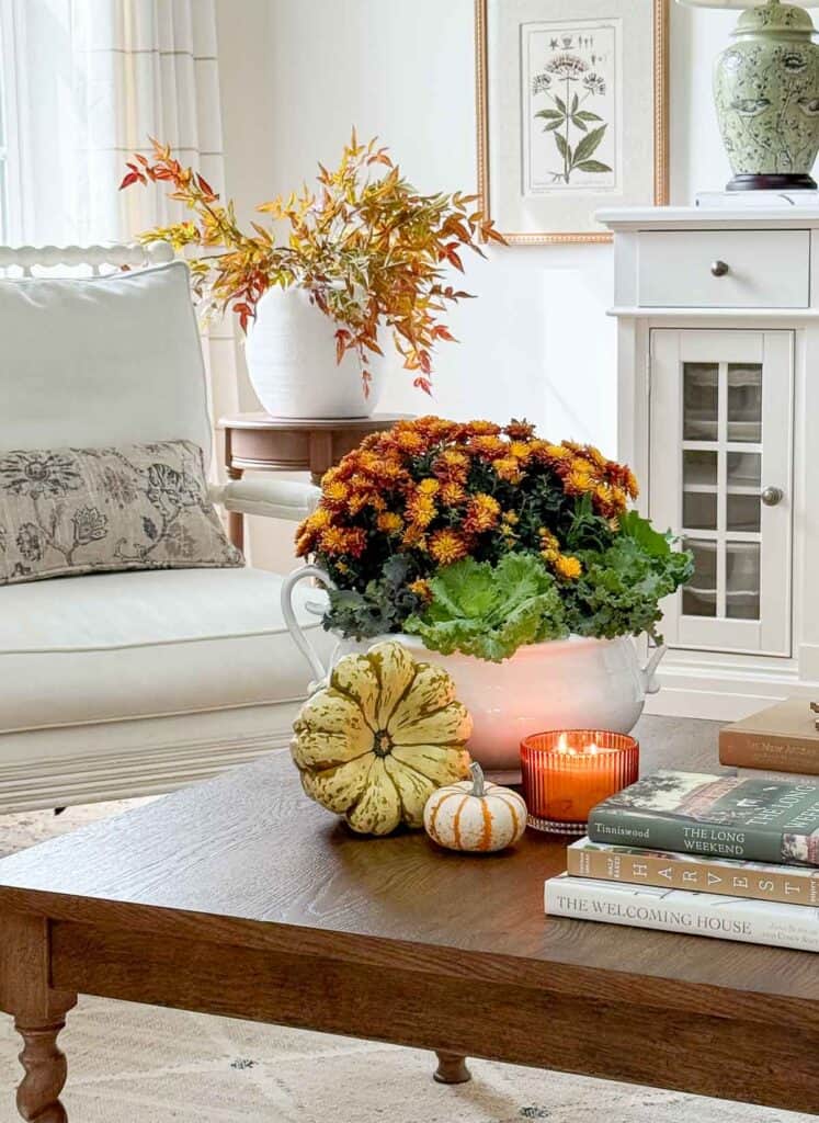 Fall coffee table centerpiece with mums in a white tureen, small pumpkins, and a glowing candle.