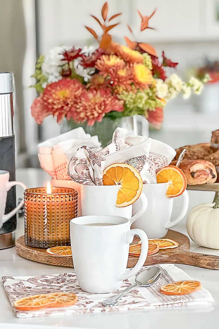 Fall coffee bar setup in a kitchen with white mugs, dried orange slices, a fall candle, and a vase of autumn flowers.