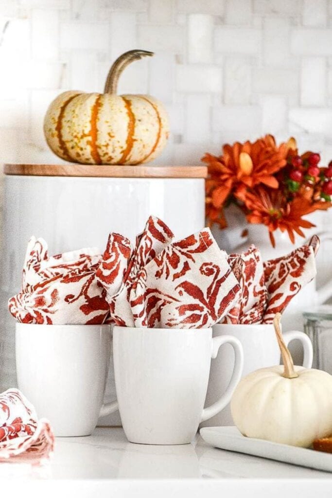 White mugs on a fall coffee bar filled with patterned red napkins, small pumpkins, and fall flowers in the background.